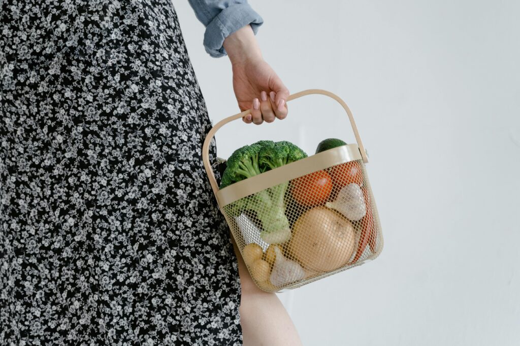 A person holding a basket with fresh vegetables like broccoli and garlic.